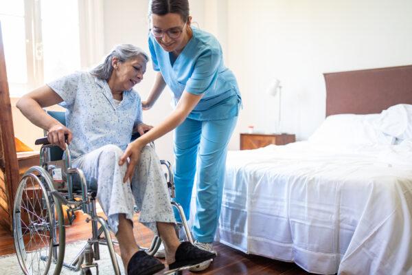 Female nurse helping senior woman to get up from wheelchair in bedroom of nursing home