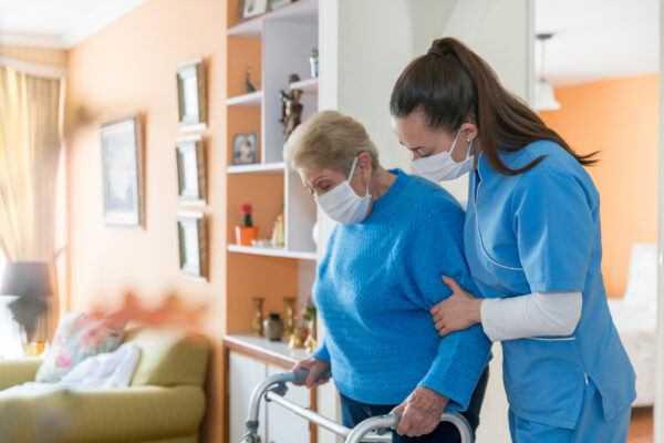 Home caregiver wearing a facemask while assisting a senior woman walking with a mobility walker during the COVID-19 pandemic - assisted living concepts